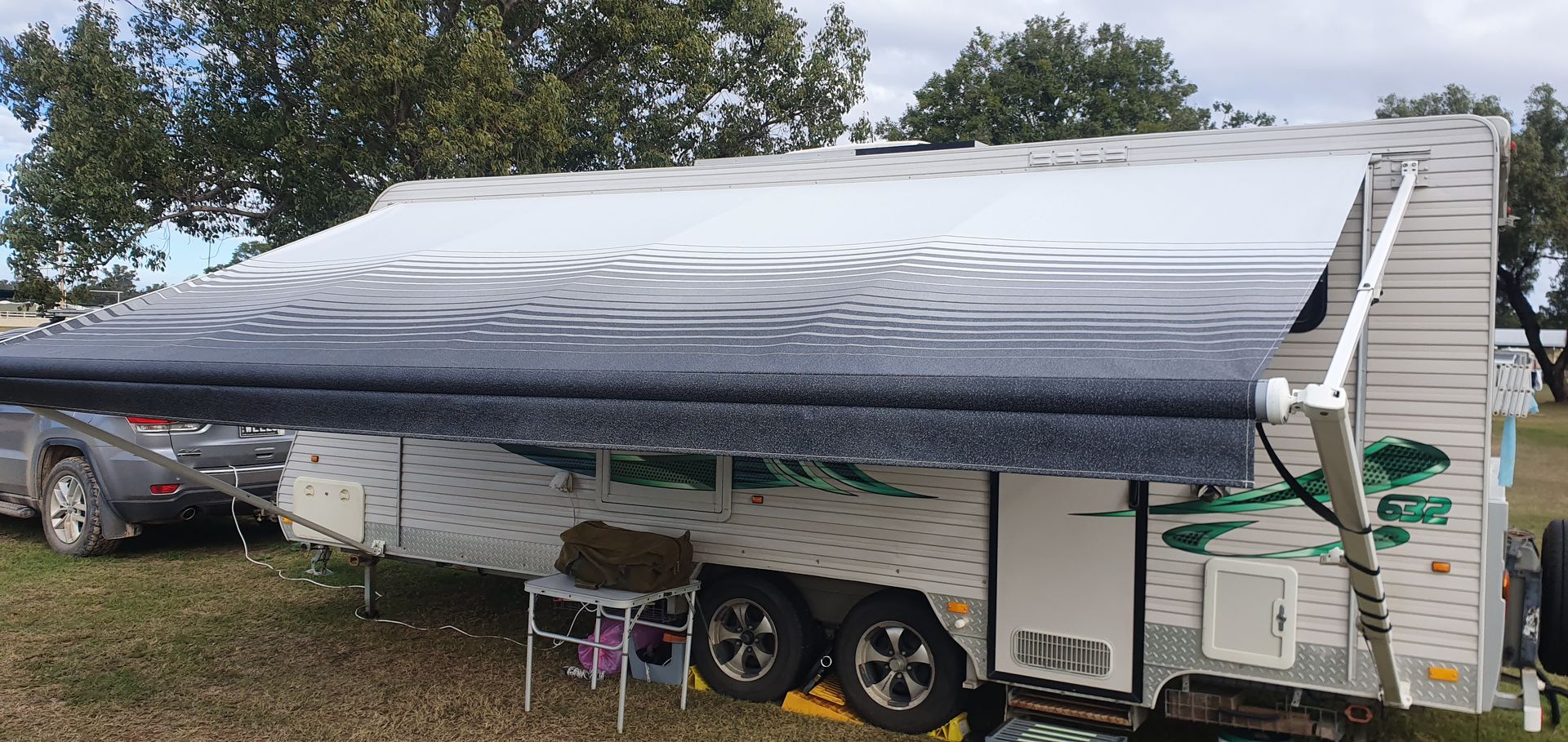 A camping trailer with its awning extended. It's parked on a grassy area, with trees in the background.