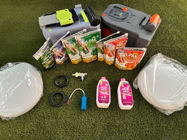 A camping toilet setup with toilet bowls, waste tanks, cleaning products, and waste treatment pouches, on green turf.