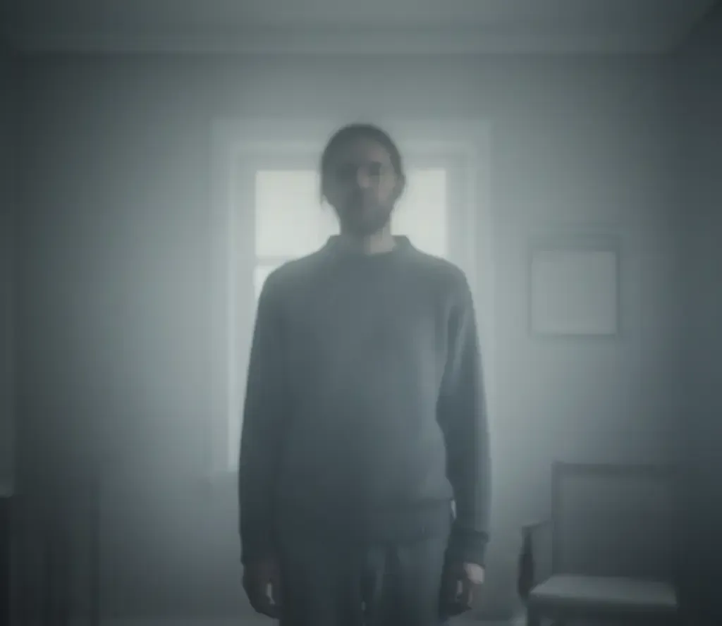 Man stands in a dimly lit room, facing forward, with a window and chair visible.