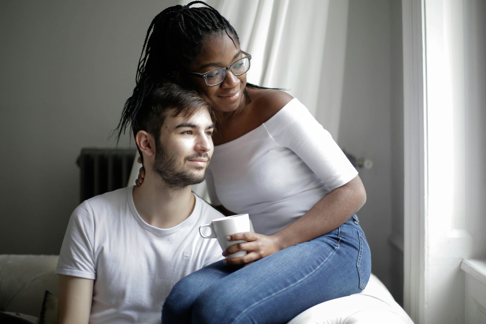 Woman in jeans and off-shoulder top hugs man, both gazing out a window with a warm, intimate expression.