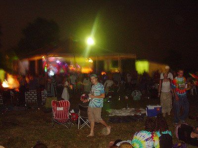 A group of people are gathered in front of a lighted stage and tent at night.
