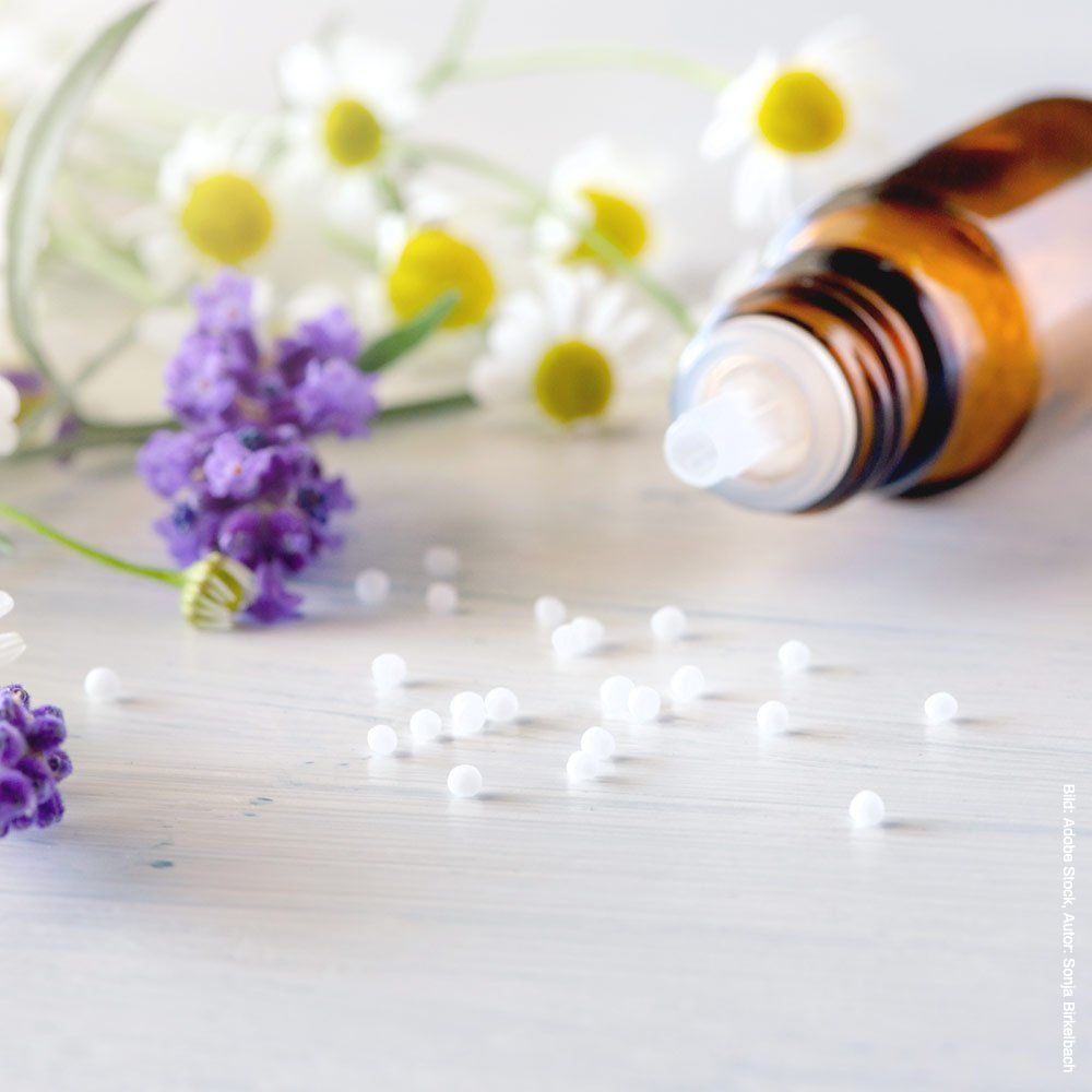 Auf einem Tisch steht neben Blumen eine Flasche Homöopathie.
