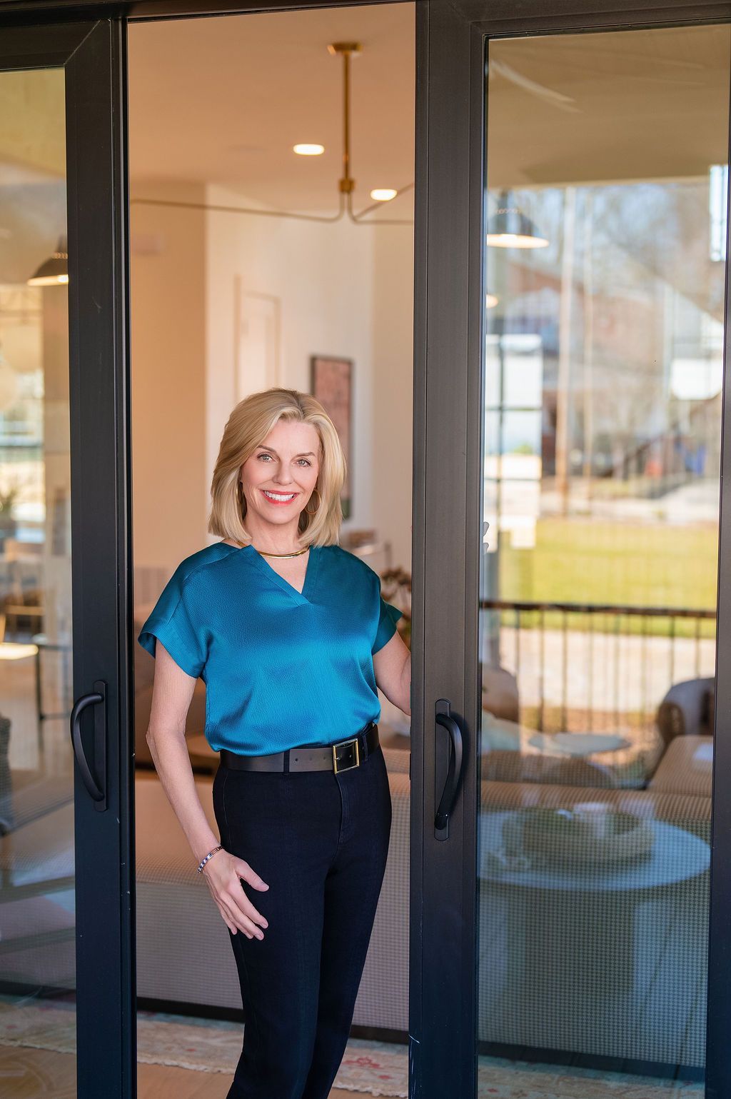 Woman standing in a doorway, smiling in a blue blouse and black pants.