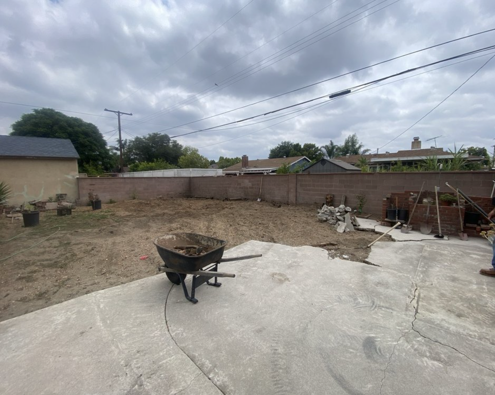 Concrete yard with wheelbarrow, dirt pile, and low brick wall under cloudy sky.