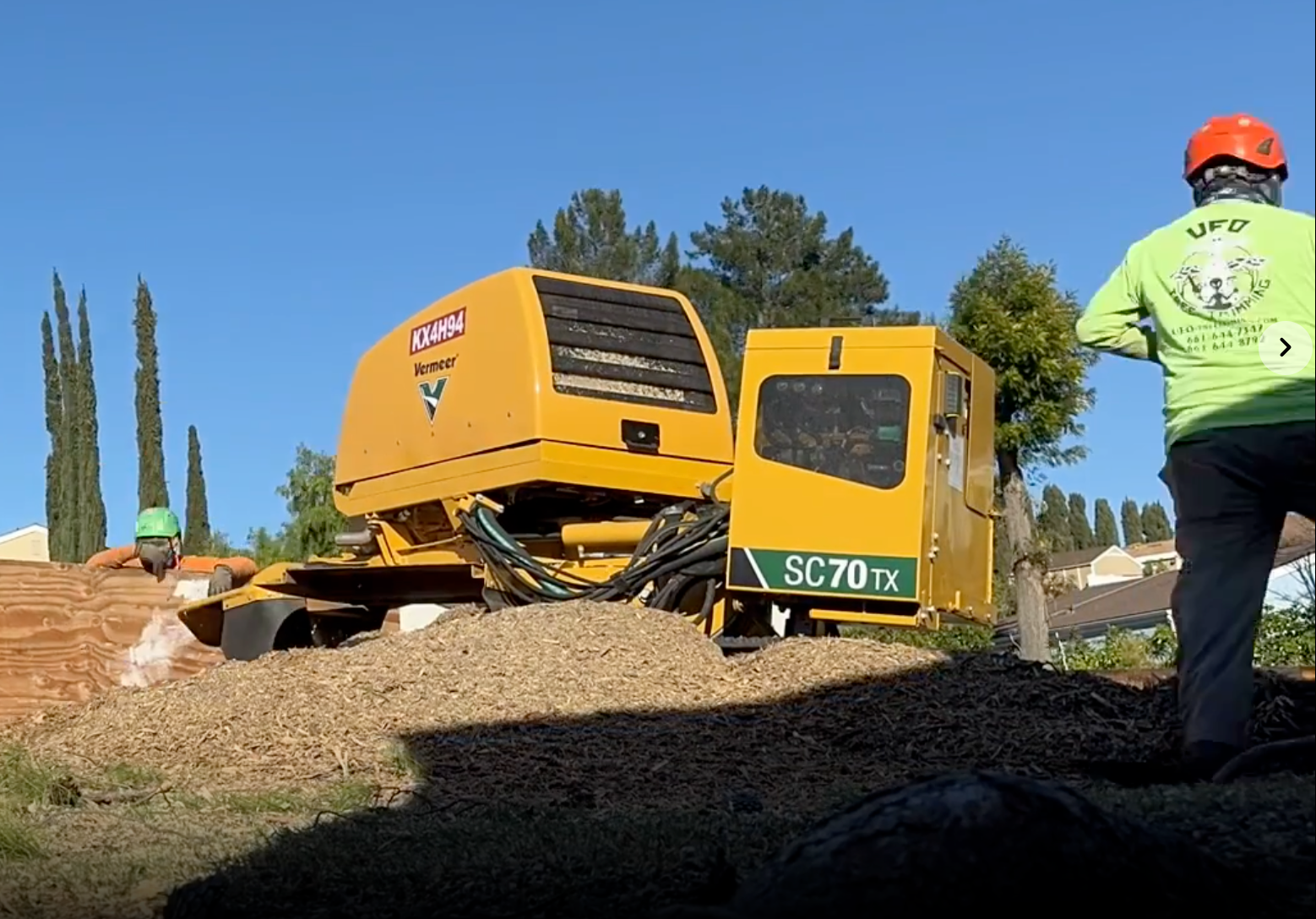 Yellow excavator on a dirt slope beside a worker in a lime shirt and orange hard hat under a blue sky
