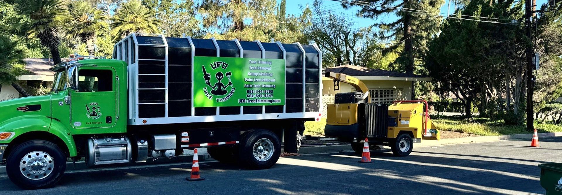 Green utility truck lifting a yellow equipment trailer on a tree-lined street