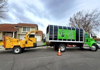 A green truck with a tree chipper attached to it is parked in front of a house.