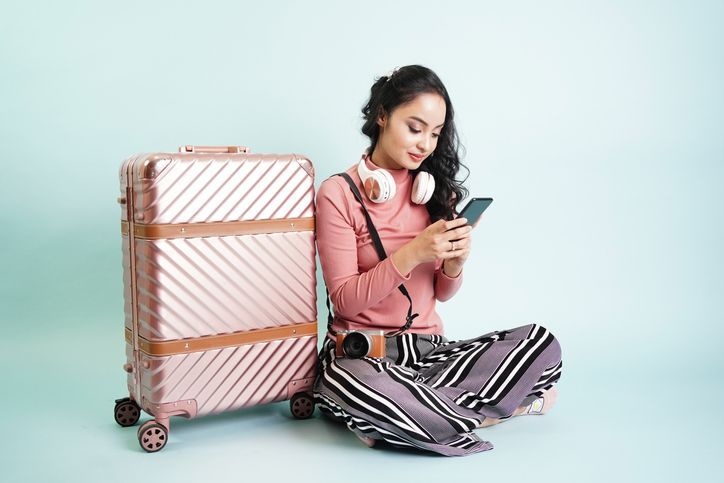 Una donna è seduta sul pavimento accanto a una valigia e guarda il suo telefono.