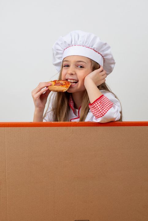 Una bambina con un cappello da cuoco sta mangiando una fetta di pizza.