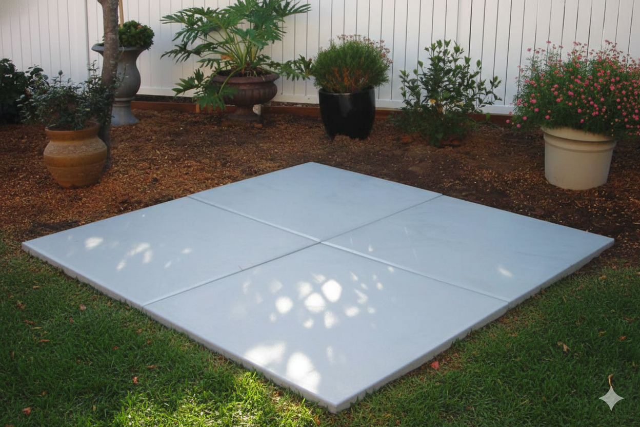 Square concrete patio on grass and gravel in a backyard, with potted plants and white fence.