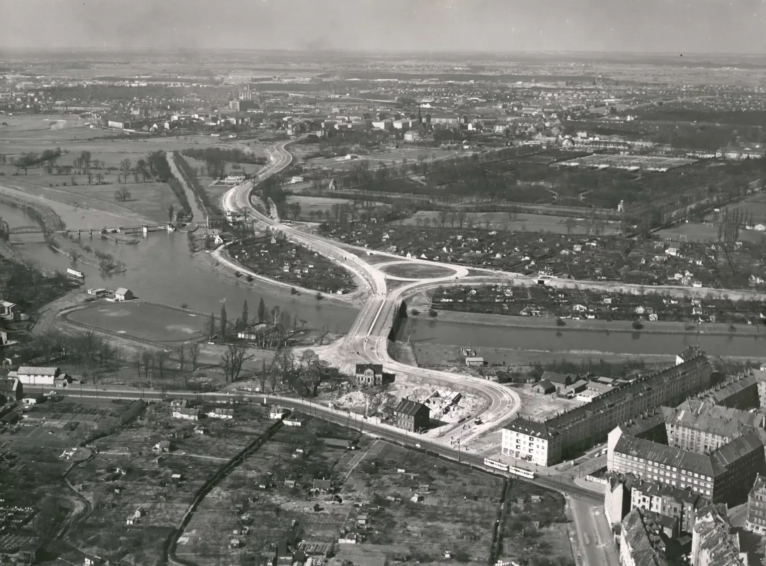 Luftbild von Luftreisedienst Niedersachsen GmbH., Hann., Frühjahr 1958: Blick auf die Leine mit Schwanenburgbrücke Westschnellweg kurz vor der Eröffnung am 1.4.1958, Schwanenburgkreisel und die Stadtteile Limmer (Leine-Verbindungs-Kanal und Leinewehr), Linden-Nord (Limmerstraße, Steigerthalstraße), Herrenhausen (Großer Garten Herrenhausen, Heizkraftwerk, Schleuse zum Ernst-August-Kanal) und Nordstadt (Wasserkunst, Westschnellweg, Schwanenburgkreisel, Bremer Damm). Ein Haus auf der Trasse hindert noch den Weiterbau der Westtangente (Westschnellweg) nach Süden zum Deisterplatz. Auf der Höhe der Straßenbahn im Vordergrund wird drei Jahre später auf dem Kleingartengelände das Freizeitheim Linden eröffnet. Bildarchiv HMH. Scan vom Foto 21×29 cm. Freigegeben durch das Historische Museum Hannover.
