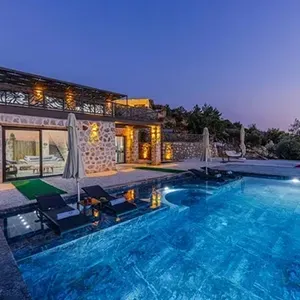 Luxury villa pool at dusk with blue water, stone facade, lounge chairs, and lit outdoor patio