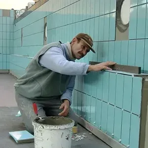 Worker tiling a wall with light blue tiles, using a trowel beside a bucket of mortar.