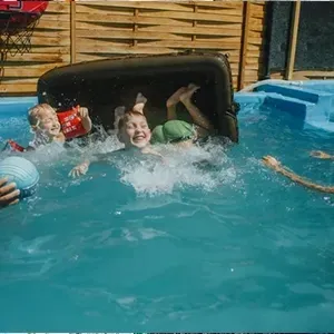 Two people splashing in a backyard pool beside an overturned black object, with blue water and wooden fencing.