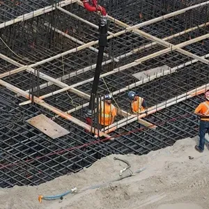 Construction workers in orange vests on a rebar grid, lifting a black hose at a concrete site