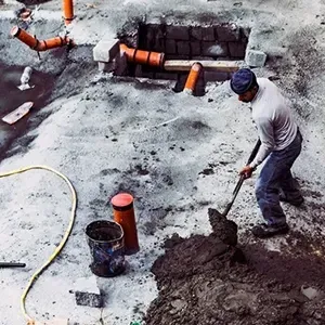 Worker shoveling wet soil at a construction site with exposed orange pipes and concrete foundation.