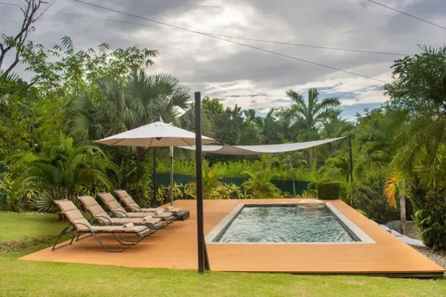 Backyard pool with loungers, umbrella, and lush tropical greenery under cloudy skies