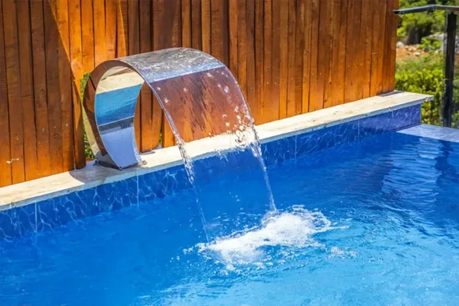 Curved stainless steel pool fountain pouring water into a blue swimming pool beside a wooden wall