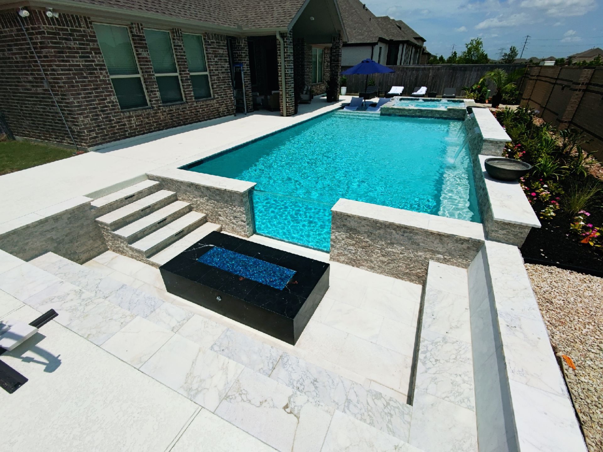 Backyard with a swimming pool, patio, and brick house surrounded by trees and shrubs.