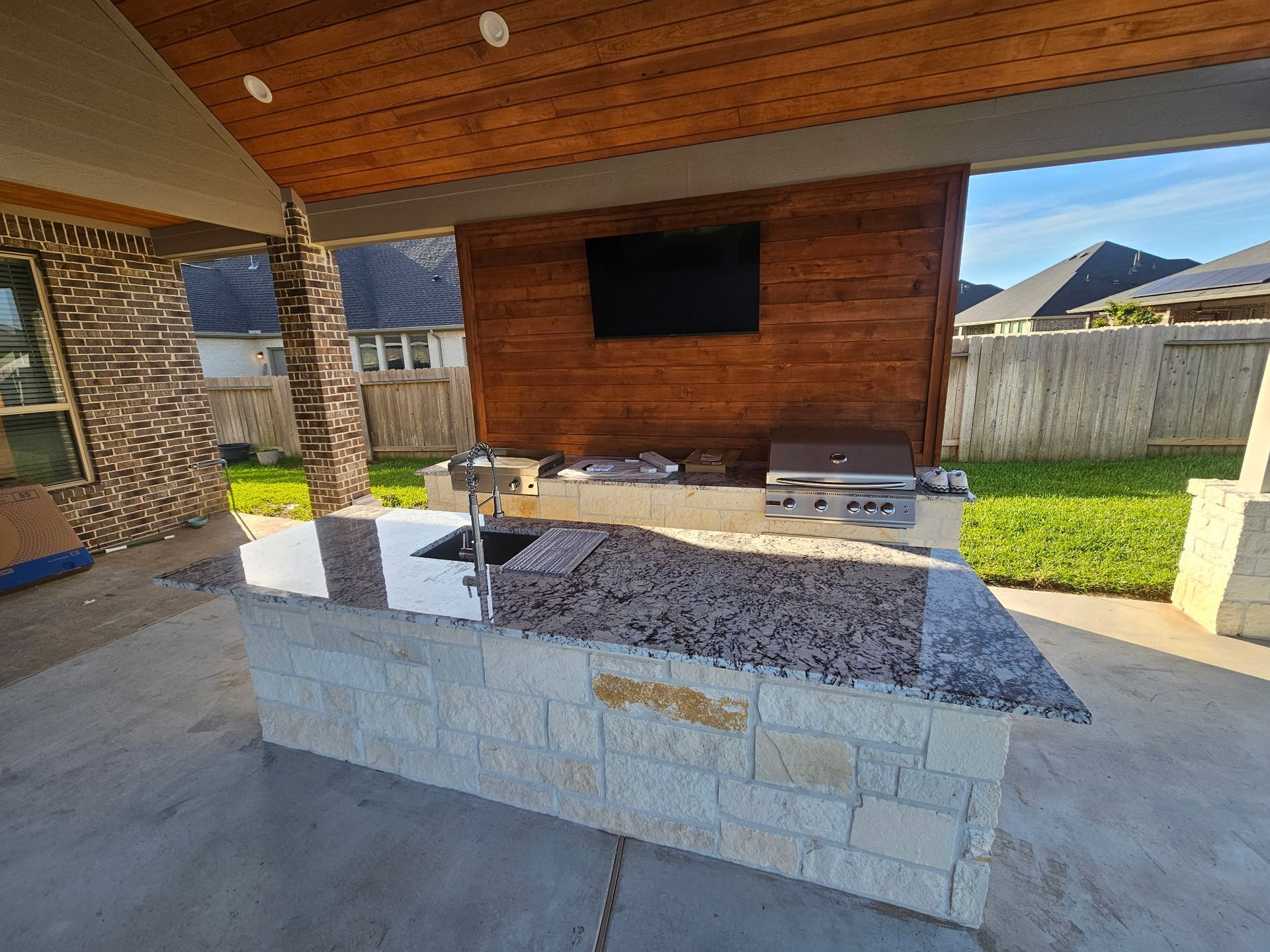 Outdoor kitchen under a covered patio with stone counter, built-in grill, and mounted TV