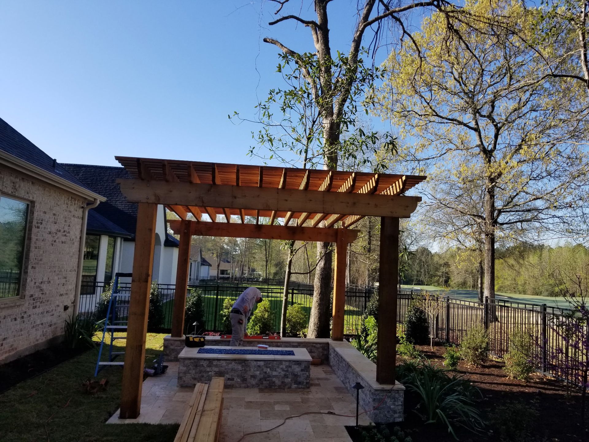 Wooden pergola with bench and stone fire pit in a sunny backyard garden