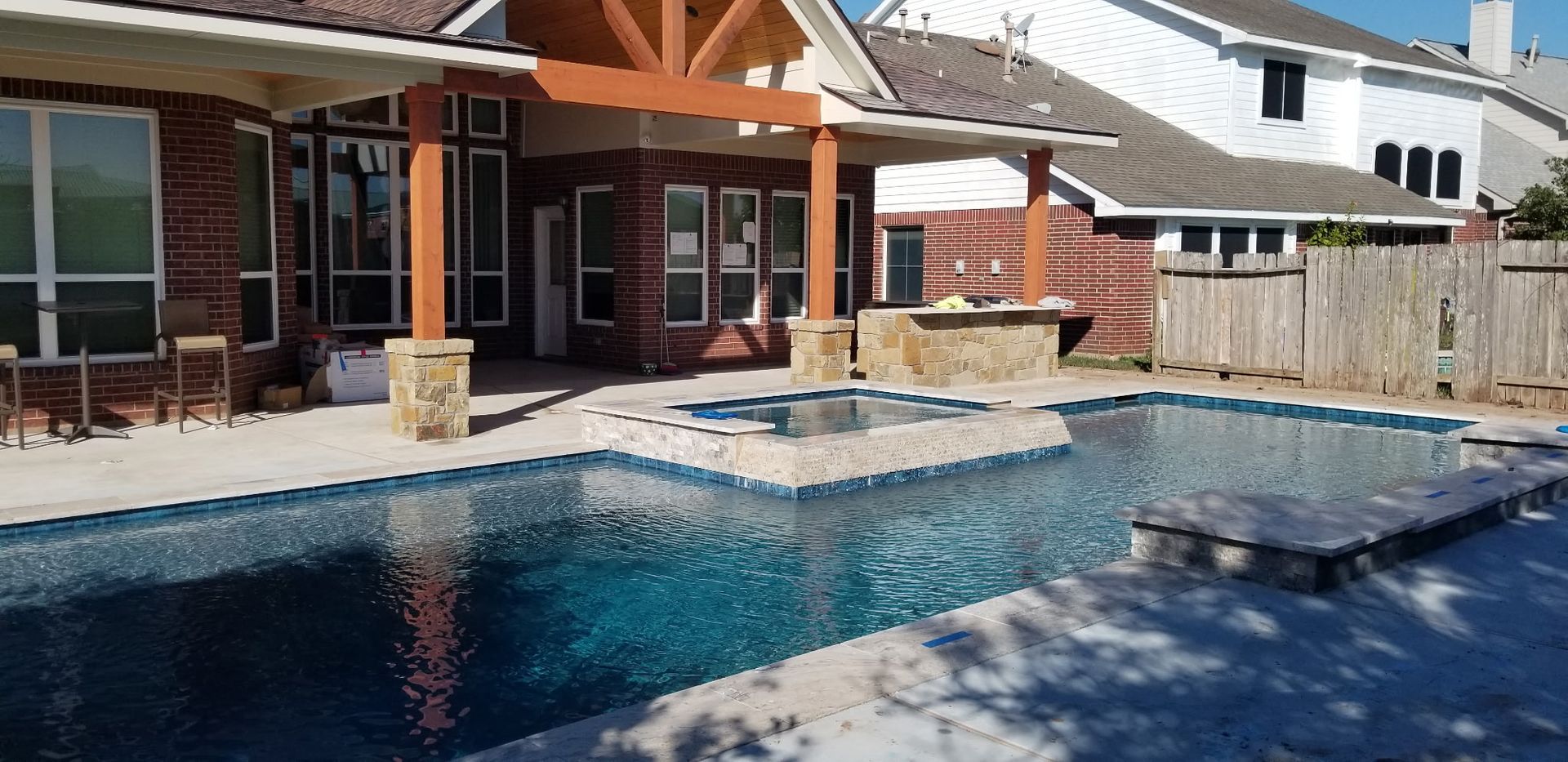 Backyard pool beside a covered patio with stone trim and neighboring homes in the background