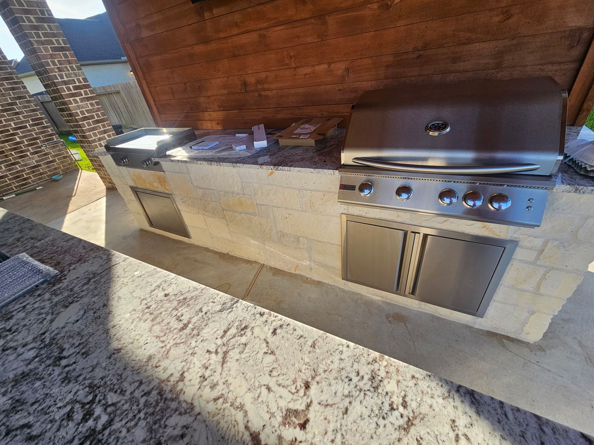 Outdoor stone-built grill and countertop under a wooden pergola, with stainless steel grill and cabinets.