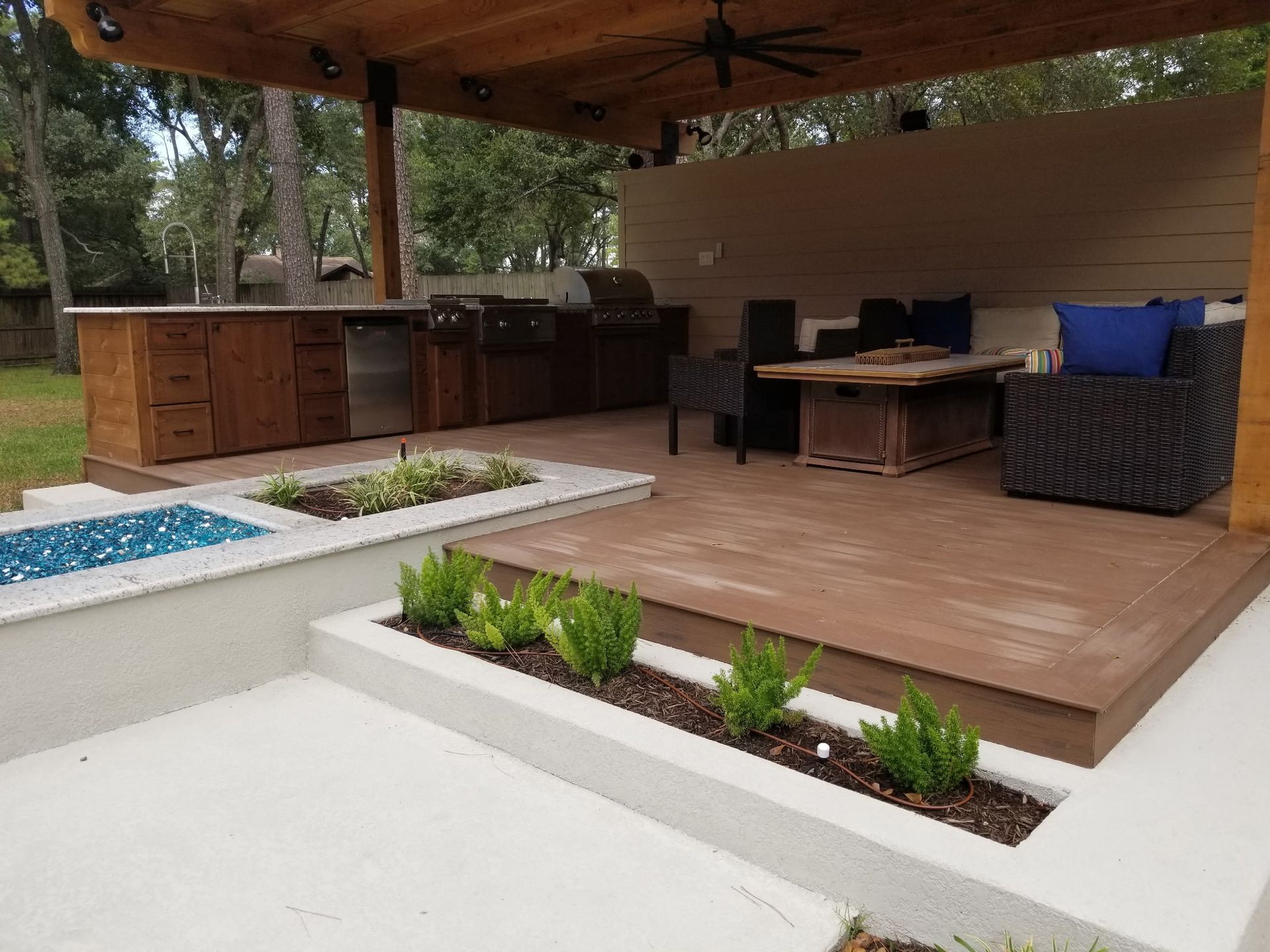 Covered backyard patio with wooden deck, outdoor kitchen, seating, and raised planter beds.