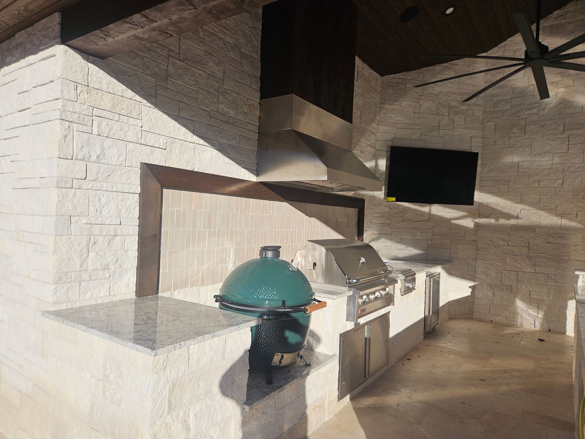 Outdoor brick patio with a green kamado grill, counter space, and a mounted TV under a covered roof