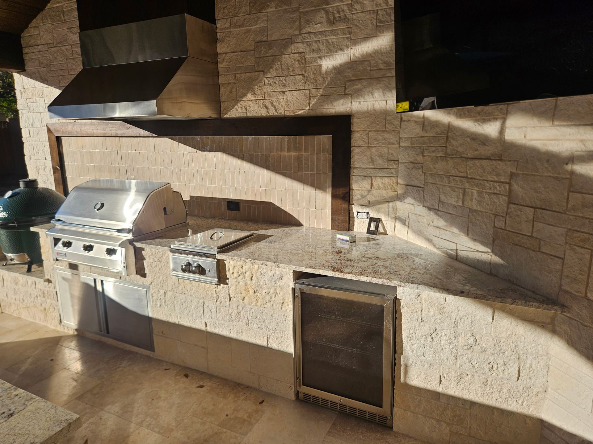 Outdoor stone kitchen with stainless steel grill, sink, and cabinets in sunlight
