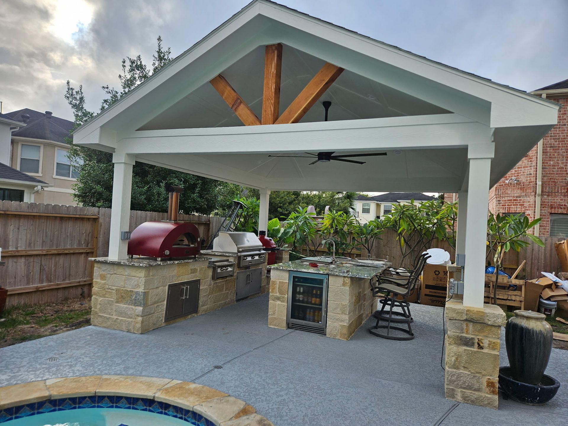 Covered backyard patio with stone bar, red grill, fan, and pool edge in front