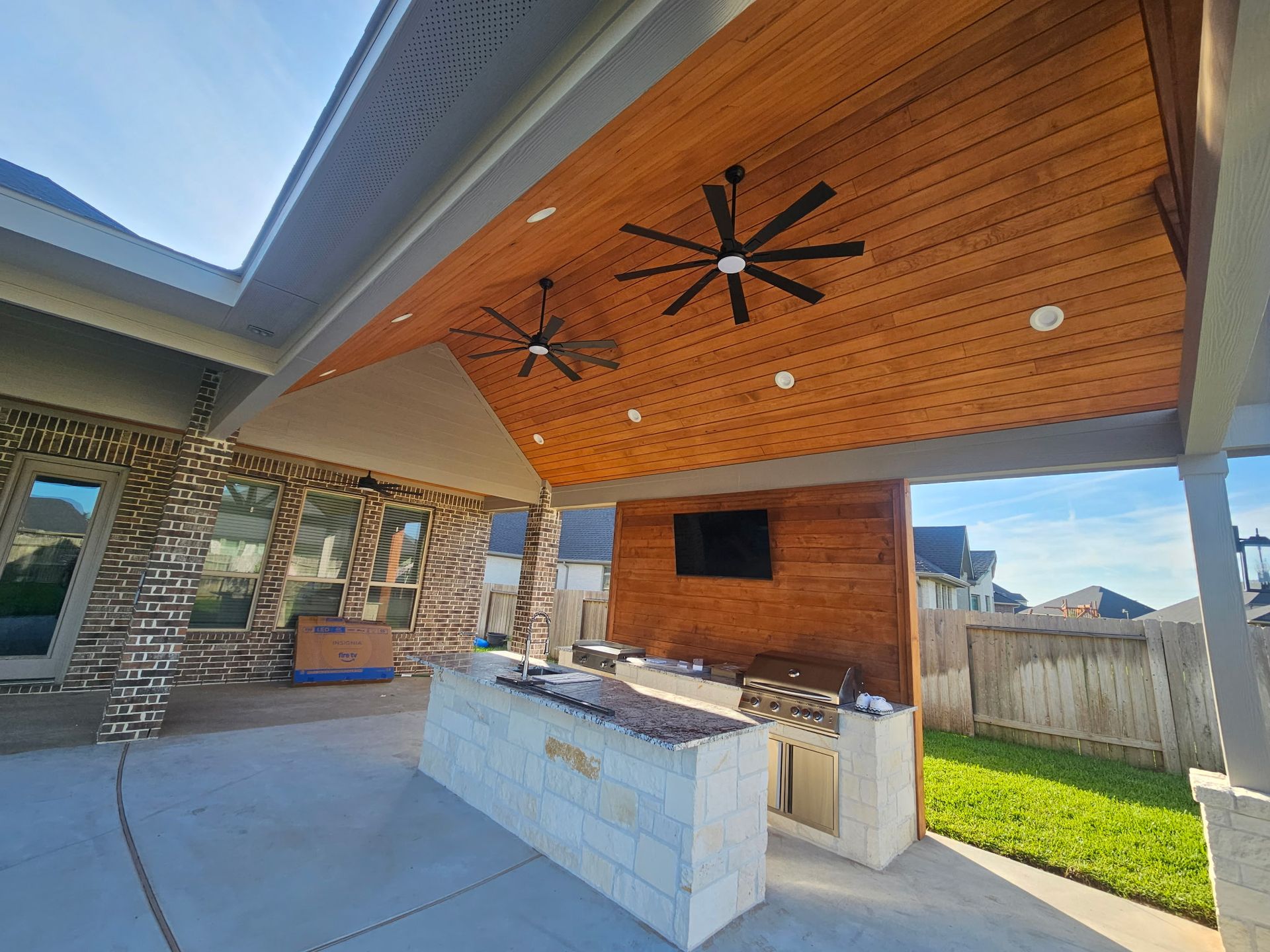 Covered outdoor patio with wood سقف, ceiling fans, built-in grill, and stone counter beside a grassy yard