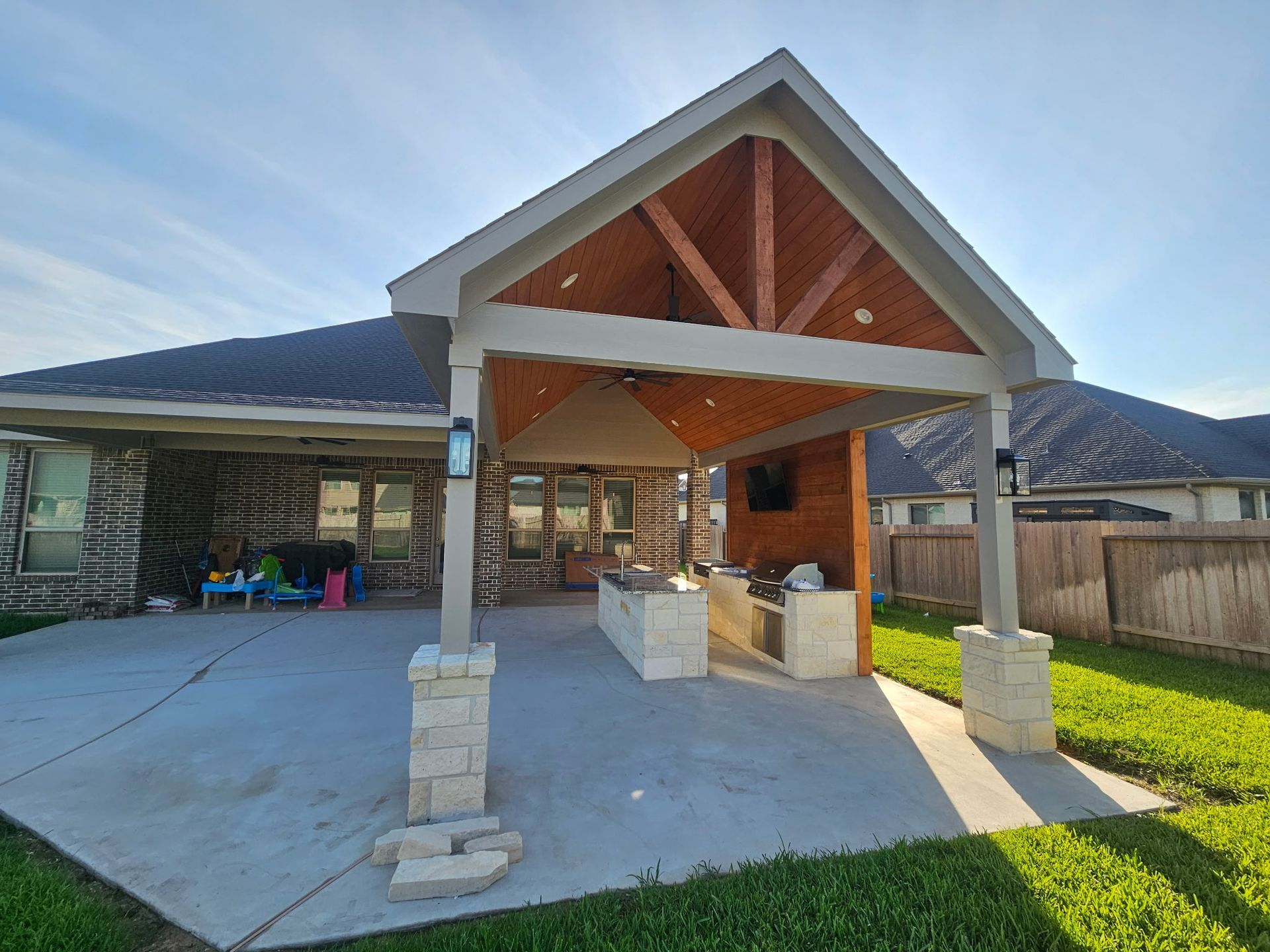 Covered backyard patio with stone columns, wood gable roof, and open concrete seating area in sunlight