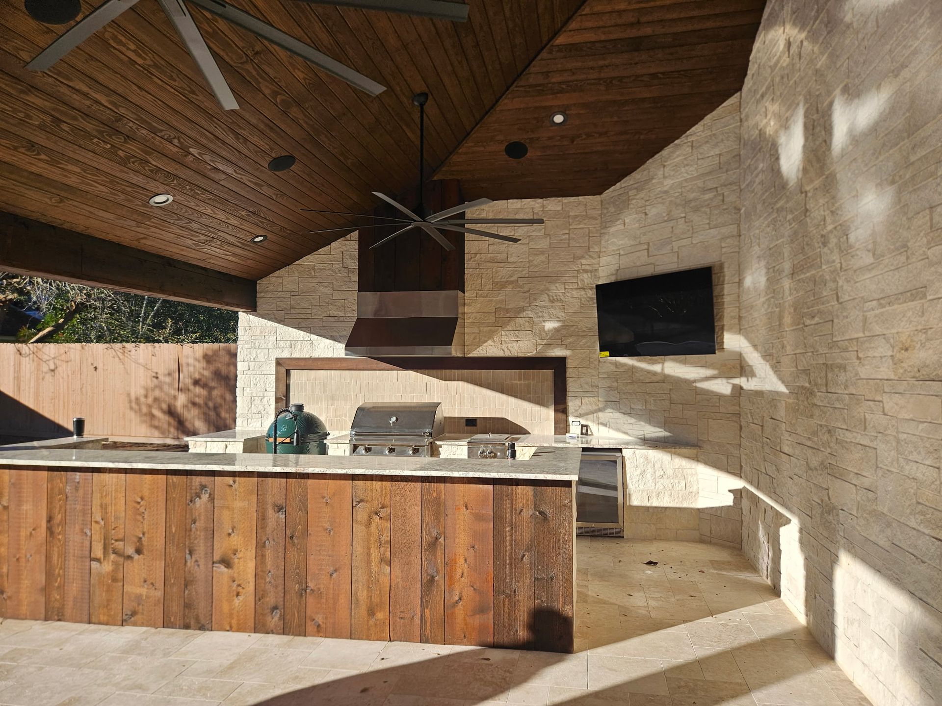 Outdoor kitchen with wood-paneled island, stone walls, and vaulted wooden ceiling under a covered patio