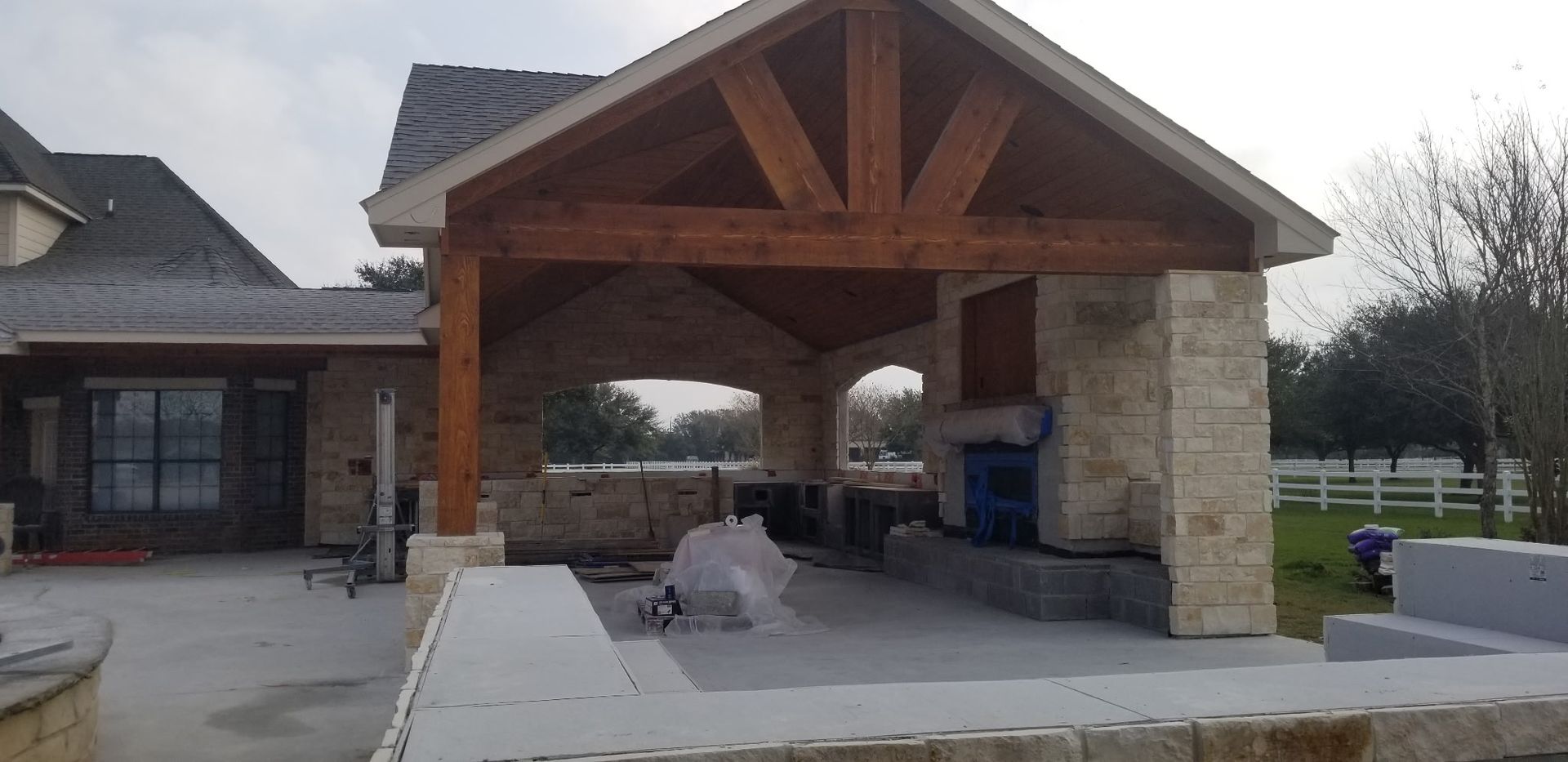 Stone-and-wood pavilion with picnic tables at a park, with a building on the left and open field in back.