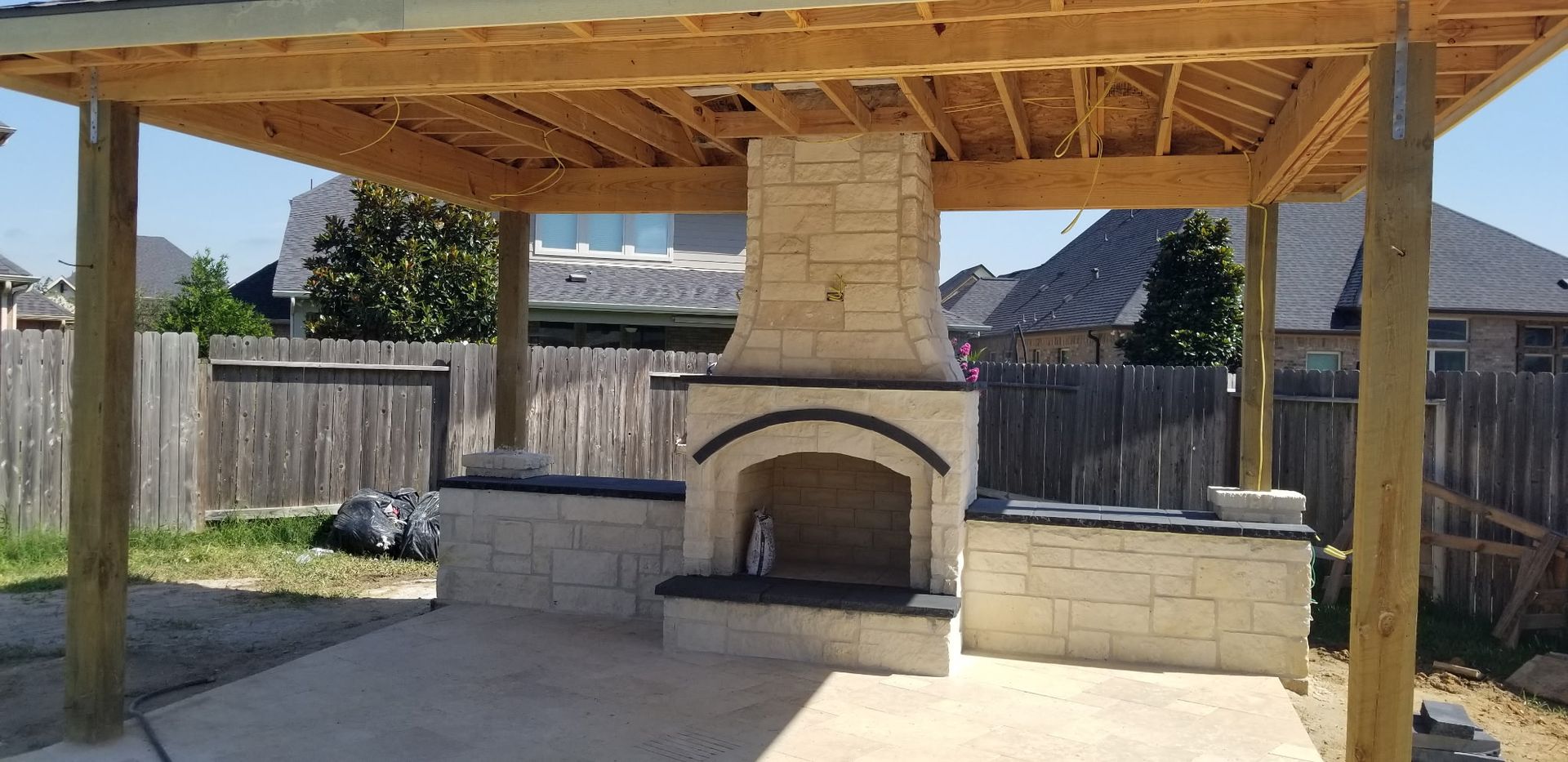 Outdoor stone fireplace and grill under a wooden pavilion in a fenced backyard