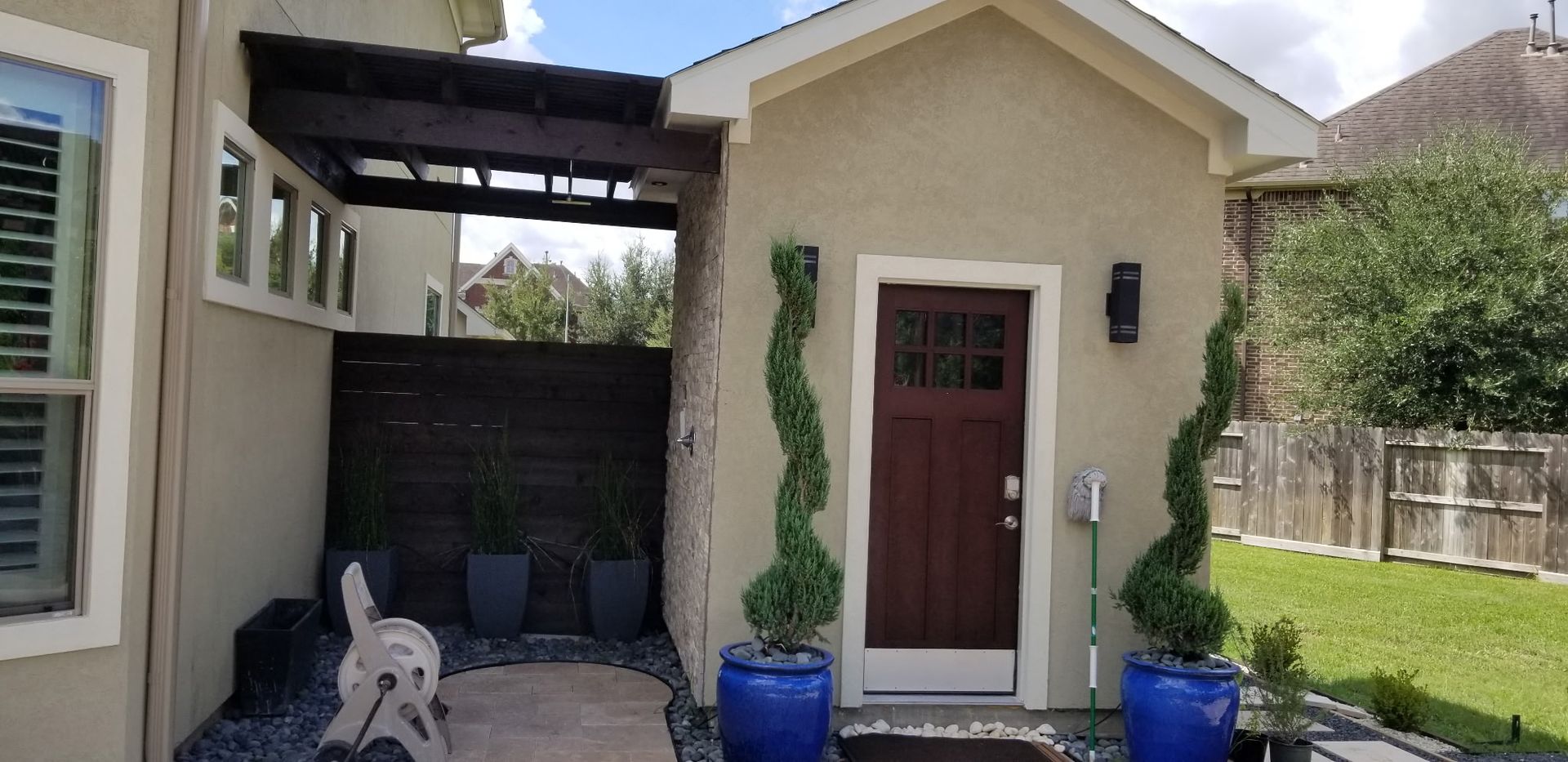 Small beige house entry with red door, blue planters, and a dog on the patio under a pergola