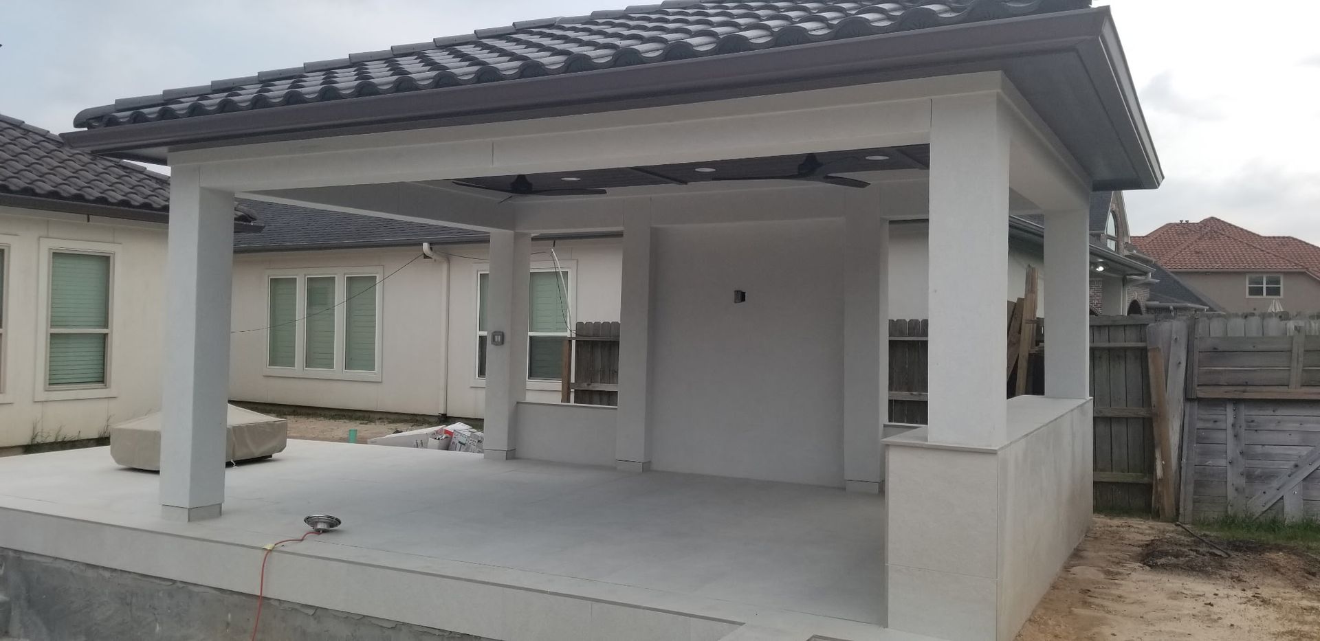 Covered backyard patio with white columns, concrete floor, and a built-in grill area next to a house.