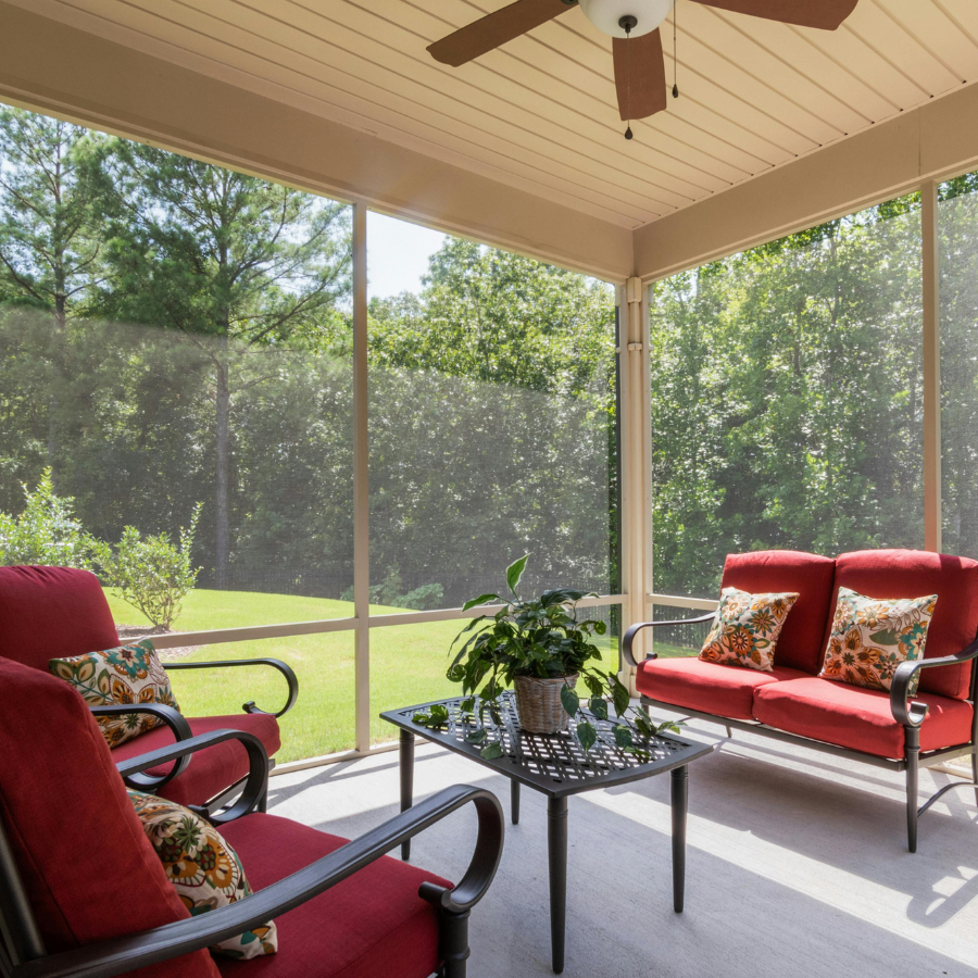 Screened porch with red cushioned chairs, glass table, and potted plant overlooking a green yard.