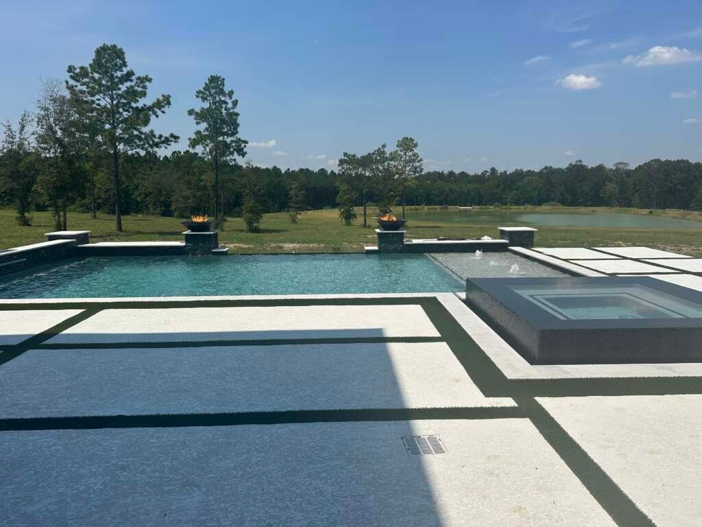 Modern backyard infinity pool with hot tub overlooking a grassy landscape and trees under a blue sky