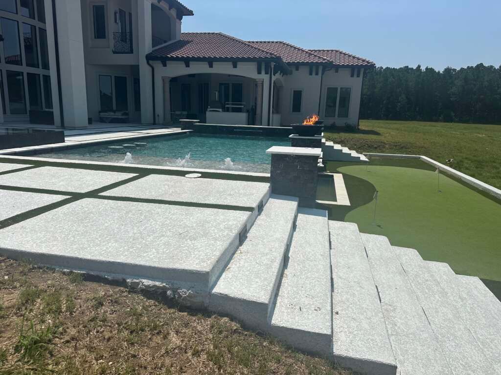 Luxury backyard pool with white stone patio, steps, and a green lawn beside a large house.