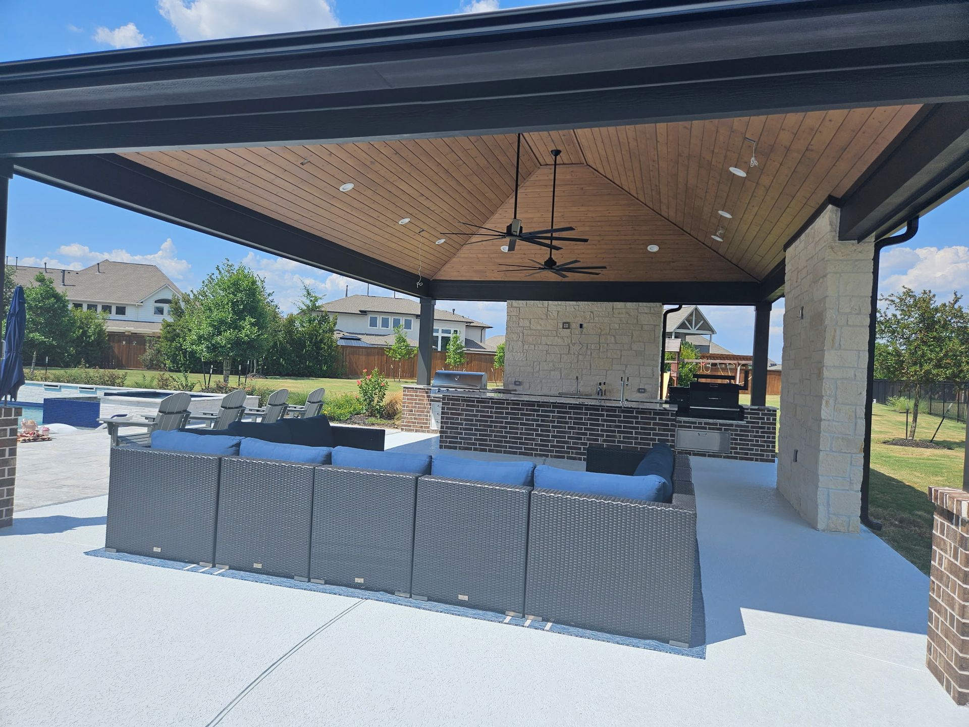 Covered outdoor patio with wicker seating, stone fireplace, and ceiling fans on a sunny day
