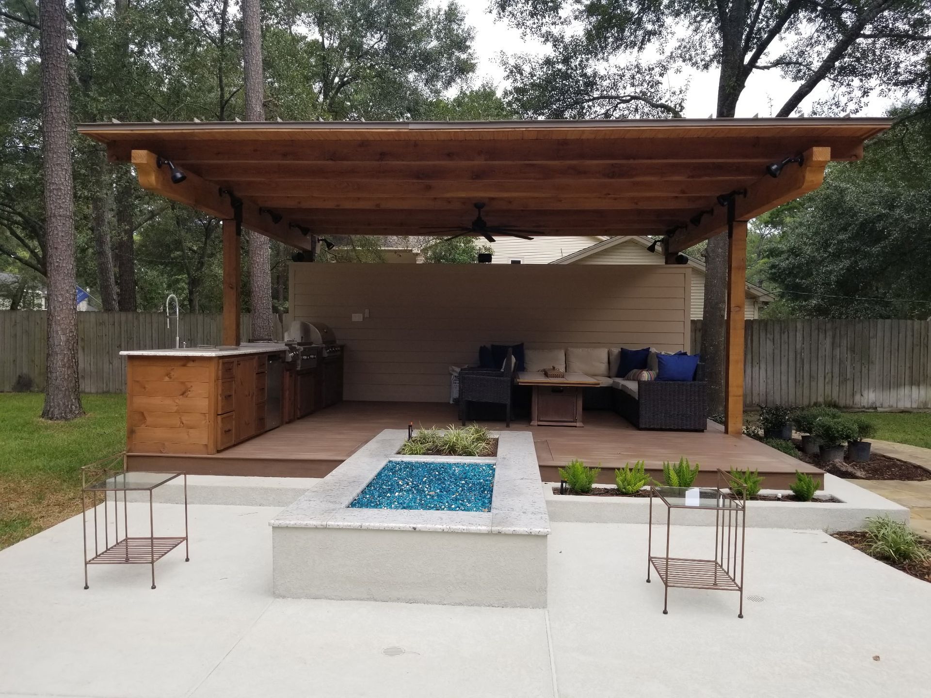 Covered backyard patio with wood roof, built-in seating, and a central white fire pit table with blue glass.