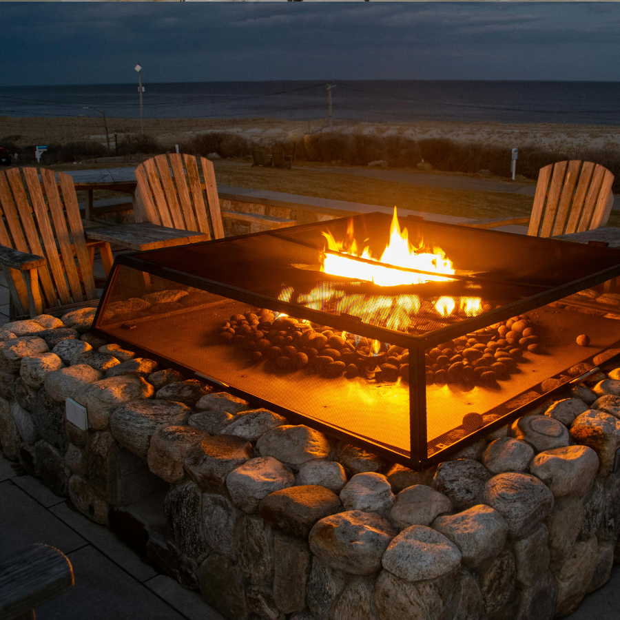 Stone fire pit with burning flames, surrounded by wooden chairs on a beach at dusk.