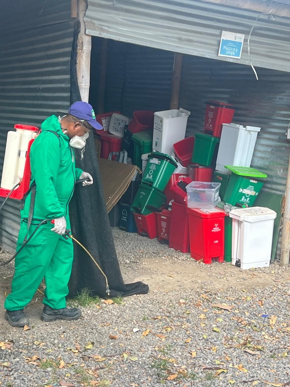 Persona con traje protector verde rociando una estructura llena de contenedores, posiblemente para saneamiento.