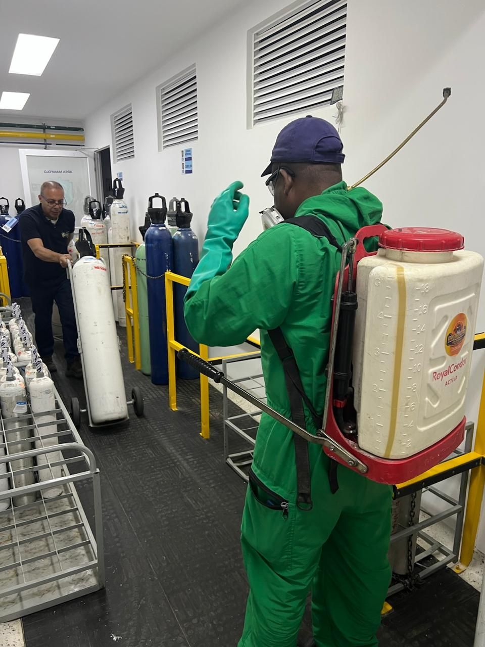 Una persona con traje protector verde rocía desinfectante cerca de cilindros de gas en un entorno industrial.