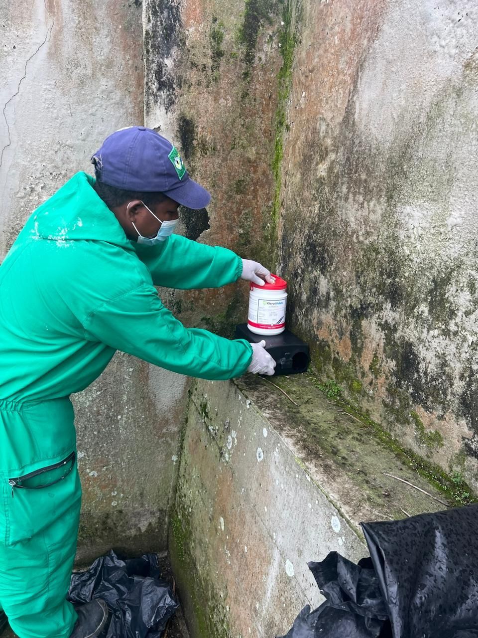 Una persona con traje de protección verde coloca un contenedor en una repisa. Al fondo, un muro de hormigón.