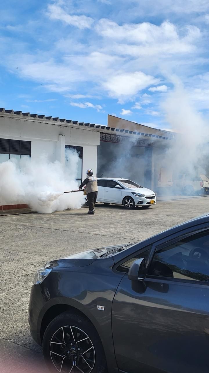 Persona rociando niebla blanca cerca de un automóvil y un edificio blancos; automóvil gris en primer plano; entorno soleado al aire libre.