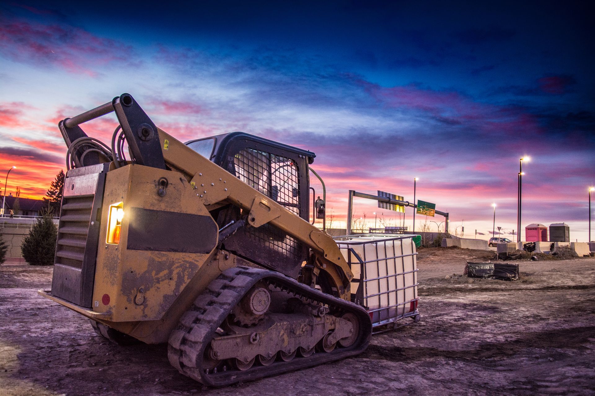 Tracked skid steer loader on a construction site at dusk with colorful sky and street lights. Tracked skid steer loader on a construction site at dusk with colorful sky and street lights.