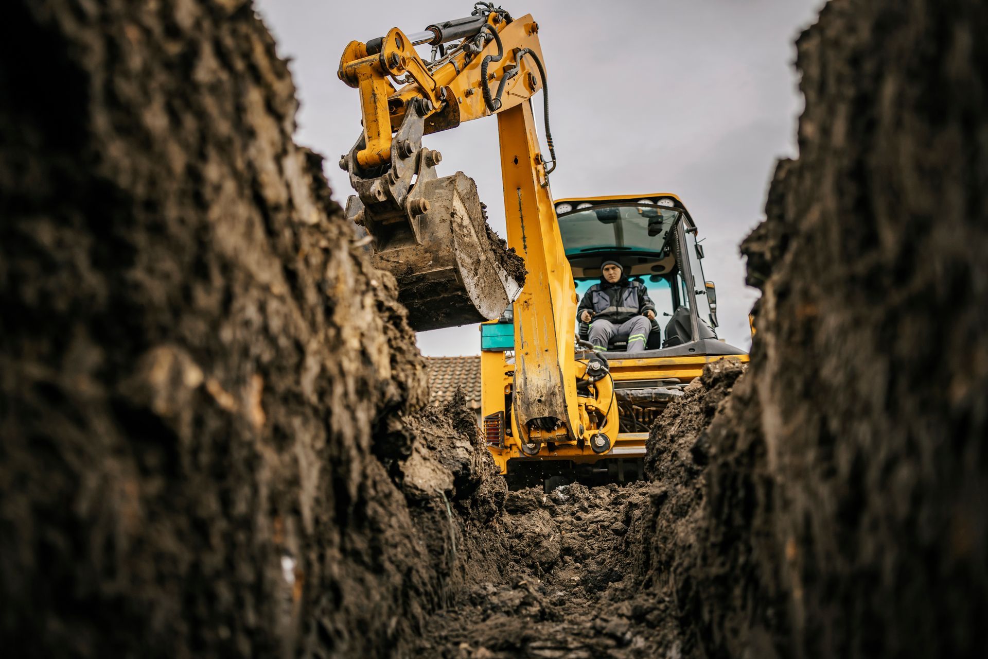 Yellow excavator digging a trench; operator in the cab. Cloudy sky in the background. Yellow excavator digging a trench; operator in the cab. Cloudy sky in the background.